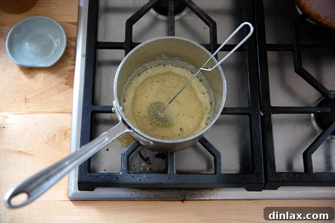 A golden-brown roux in a small saucepan on the stovetop, being continuously whisked to ensure a smooth consistency.
