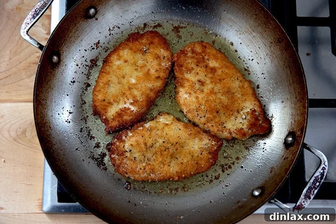 Three chicken cutlets frying in a large skillet on the stovetop.