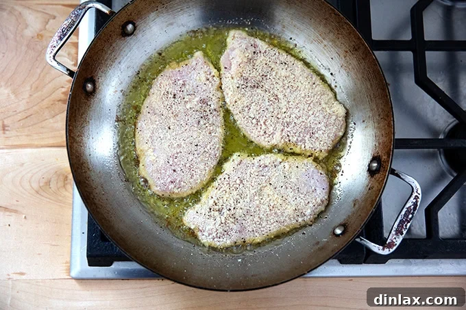 Three chicken cutlets frying in a large skillet on the stovetop.