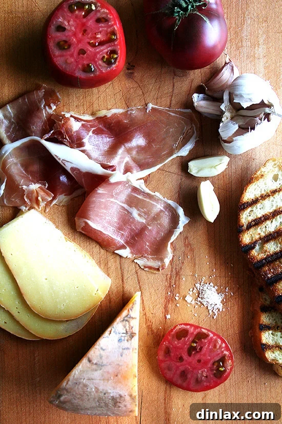 Various ingredients laid out for making the ultimate Spanish sandwich, including rustic bread, garlic, a ripe tomato, a bottle of olive oil, cured ham, and Spanish cheese.