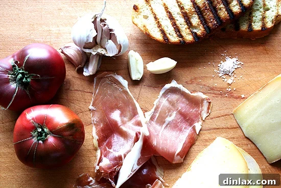 Essential ingredients for Pan con Tomate laid out on a table: a ripe red tomato, a head of garlic, and a bottle of extra-virgin olive oil.
